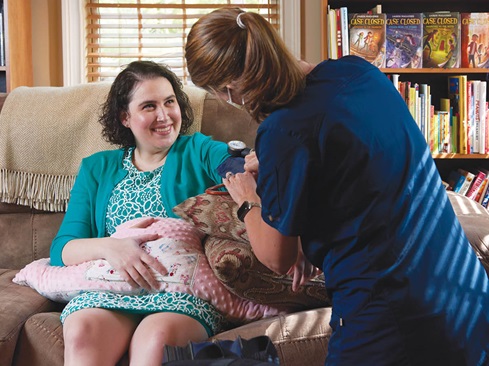 Lauren Magaziner sits on a couch at home while Erin McCarthy, RN, NP, checks her blood pressure.